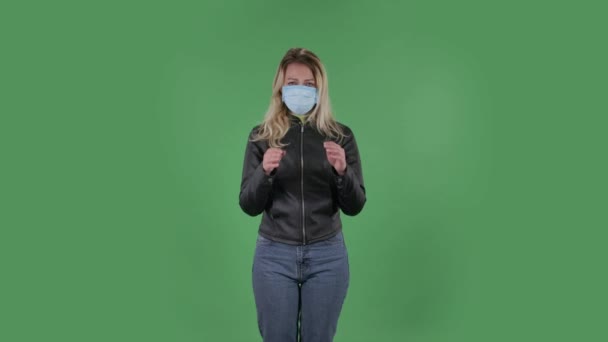 Portrait de belle jeune femme en masque médical regarde droit et prie mains jointes paume à paume. Blonde avec des cheveux amples dans une veste noire et un jean sur un écran vert dans le studio. Santé — Video