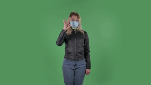 Ritratto di bella giovane donna in maschera medica sta guardando la macchina fotografica e gioiosamente facendo segno ok. Bionda con i capelli sciolti in una giacca nera e jeans su uno schermo verde in studio. Sanità — Video Stock