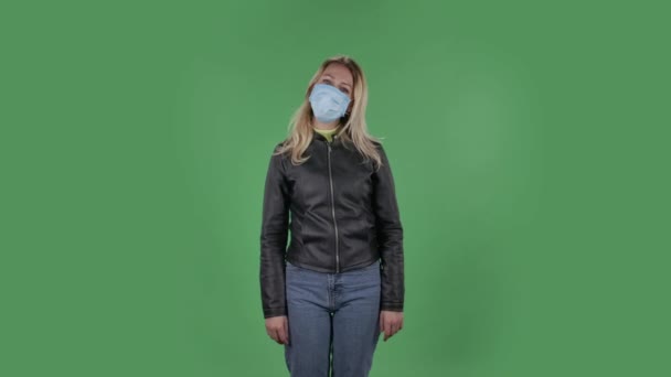 Portret van mooie jonge vrouw met medisch masker is recht kijken zucht opgelucht en het maken van teken ok. Blond met los haar in een zwart jasje en jeans op een groen scherm in de studio. Gezondheid — Stockvideo