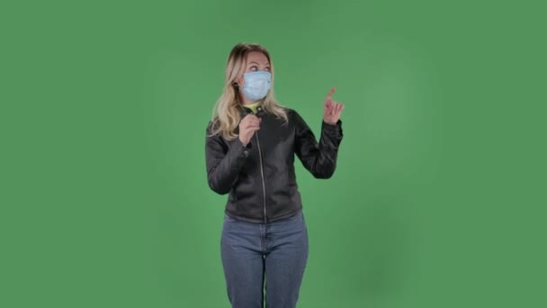 Ritratto di bella giovane donna in maschera protettiva medica guardando la macchina fotografica e indicando le dita di lato. Bionda con capelli sciolti in una giacca nera e jeans su uno schermo verde in studio — Video Stock