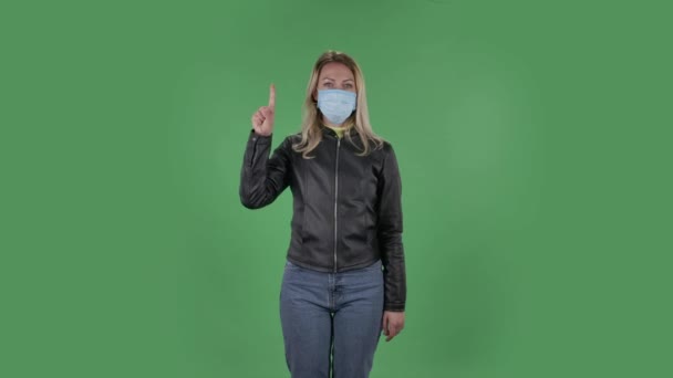 Portret van een mooie jonge vrouw met een medisch beschermend gezichtsmasker kijkt naar de camera en toont vuist, telt dan één, twee, drie, vier, vijf vingers. Blond met los haar in een zwart jasje en — Stockvideo