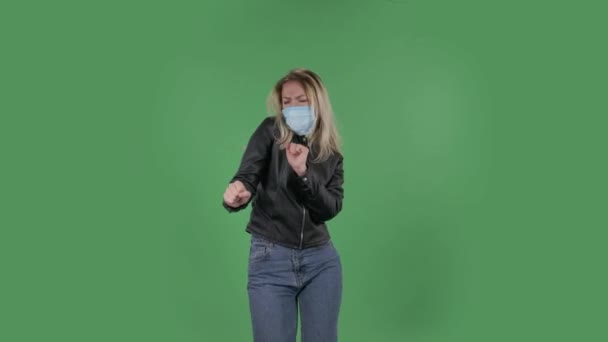 Retrato de una hermosa mujer joven en máscara médica se ve recta y está bailando divertido. Rubia con el pelo suelto en una chaqueta negra y jeans en una pantalla verde en el estudio. Protección de la salud Corona — Vídeos de Stock