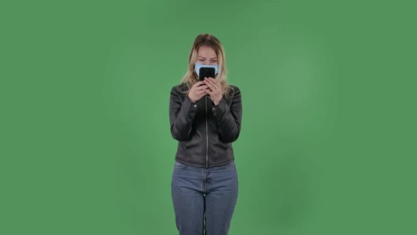 Ritratto di bella giovane donna in maschera medica sta messaggiando sul suo telefono. Bionda con i capelli sciolti in una giacca nera e jeans su uno schermo verde in studio. Protezione della salute Corona Virus Concept. — Video Stock