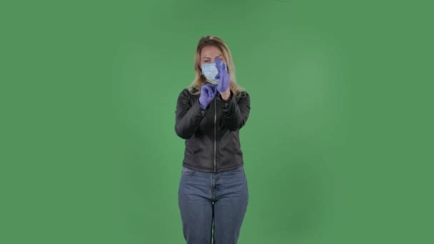 Portret van een mooie jonge vrouw met een medisch masker kijkt naar de camera en trekt rubberen handschoenen aan. Blond met los haar in een zwart jasje en jeans op een groen scherm in de studio. Gezondheidsbescherming — Stockvideo