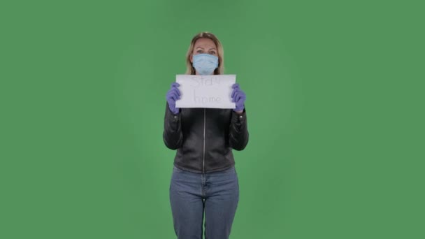 Portrait de belle jeune femme en masque médical regarde la caméra et tient une affiche avec les mots rester à la maison. Blonde aux cheveux amples dans une veste noire et un jean sur un écran vert dans le studio — Video