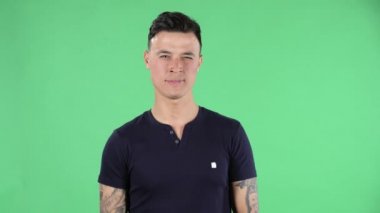 Yakışıklı bir gencin portresi el sallıyor ve jest yapıyor. Buraya gel. Siyah tişörtlü, esmer, şık saçlı, elinde dövmeleri olan, stüdyoda yeşil bir ekranda..