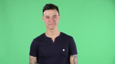 Portre genç adam neşeyle elini sallıyor ve buraya gelip göz kırpmak için bir jest gösteriyor. Siyah tişörtlü, esmer, şık saçlı, elinde dövmeleri olan, stüdyoda yeşil bir ekranda..