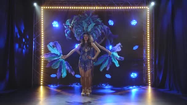 Un grupo de encantadoras mujeres jóvenes en trajes de carnaval brasileño azul, sombreros y abanicos de plumas están bailando en el estudio. Espectáculo femenino de danza en el fondo de luces de neón dinámicas. Movimiento lento. — Vídeo de stock