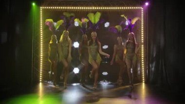 Karanlık bir sahnede sarı ışıklarla dans eden güzel bir kadın grubunun dans gösterisi. Topuklu giyen kadınların takım dansı. Yavaş çekim.