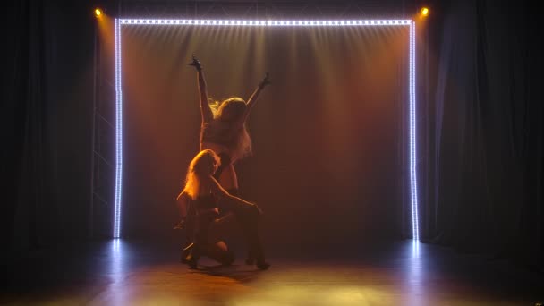 Дві молоді жінки йдуть танцюристами з довгим волоссям еротично танцювати в темній студії з поставленим світлом. Силуети струнких тіл рухаються у повільному русі . — стокове відео
