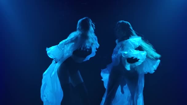 Silhouettes en gros plan de deux jeunes femmes sexy dansant en capes blanches aérées et lingerie noire. Tourné dans un studio sombre avec des néons bleus. Mouvement lent. — Video