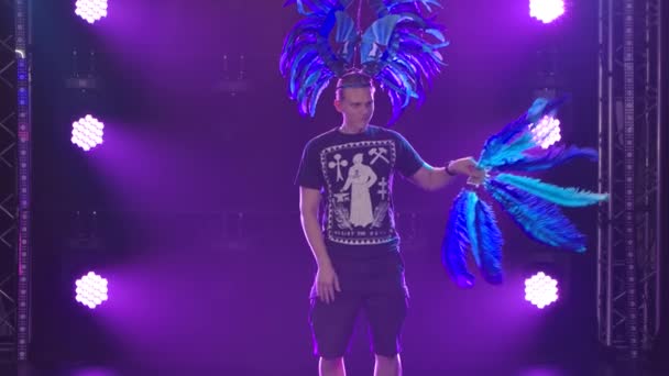 Un jeune homme blond dans une coiffure et avec un ventilateur de plumes bleues danse drôle dans le studio sur un fond de lumières violettes. Ferme là. Mouvement lent. — Video