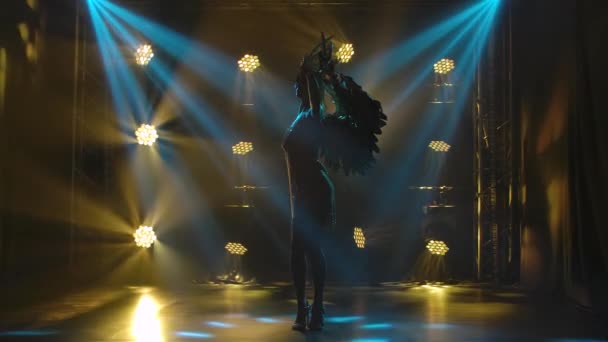 A young actress in the image of the Greek goddess Artemis poses on stage in the rays of magical light. Silhouette of a beautiful woman with golden wings and a headdress with feathers. Slow motion. — Stock Video