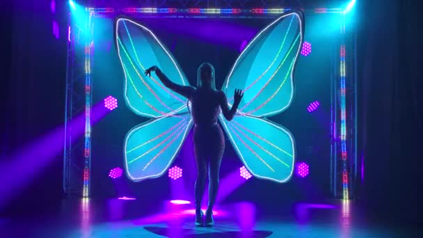 Silhouette of a slender female singer in a tight-fitting jumpsuit strewn with rhinestones and with glowing butterfly wings behind her back. Slow motion. — Stock Video