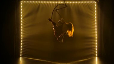 Ringde siluet hava akrobatı var. Genç bir kadın, yumuşak sarı ışıkla çevrili hava halkasındaki akrobatik elementleri sergiliyor. Yavaş çekim.