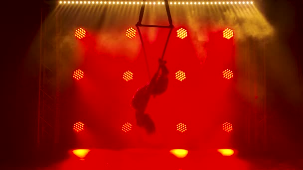 Mujer morena bonita flexible en un traje ajustado púrpura realiza elementos acrobáticos en un aro de aire. Un artista de circo giratorio silueta sobre un fondo de luces de neón rojas. Movimiento lento. — Vídeos de Stock