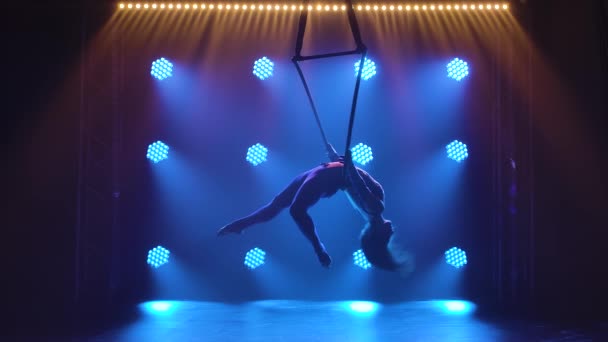 Silhouette woman aerialist performs acrobatic elements and spins in hanging aerial hoop against background of blue neon lights. Slow motion. — Stock Video