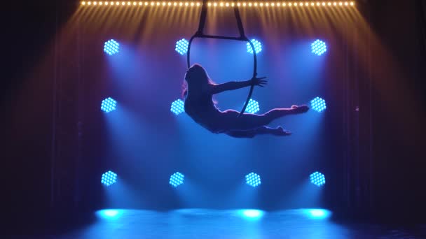 Aerialista mujer silueta realiza elementos acrobáticos y gira en aro aéreo colgante contra el fondo de las luces de neón azul. Movimiento lento. — Vídeos de Stock