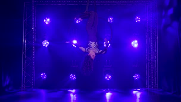 Woman aerialist performs acrobatic elements and spins in hanging aerial hoop against background of blue neon lights. Slow motion. — Stock Video