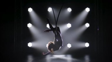 Güzel kıvırcık hava jimnastikçisi hava çemberinde elementler sergiliyor. Sahne ışıkları altında genç bir kızın zarif silueti karanlığın içinde beliriyor. Yavaş çekim.