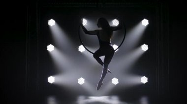 Güzel kıvırcık hava jimnastikçisi hava çemberinde elementler sergiliyor. Sahne ışıkları altında genç bir kızın zarif silueti karanlığın içinde beliriyor. Yavaş çekim.