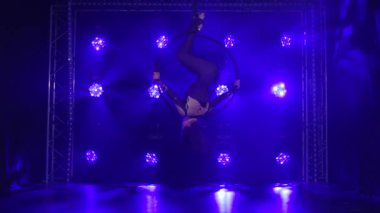Kadın ateist akrobatik elementler sergiliyor ve mavi neon ışıkların arka planına karşı asılı duran hava çemberinde dönüyor..