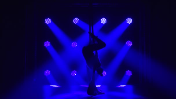 Kvinnlig antenn jämvikt gymnast utför balansering på en luft silke i en mörk studio med blå scenbelysning. Silhuett av en smal kropp. Långsamma rörelser. — Stockvideo