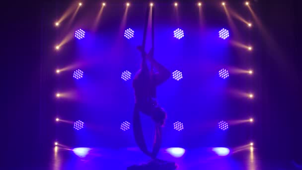 Gimnasta de equilibrio aéreo femenina que realiza el equilibrio en una seda de aire en un estudio oscuro con iluminación de escenario azul. Silueta de cuerpo delgado. Movimiento lento. — Vídeos de Stock