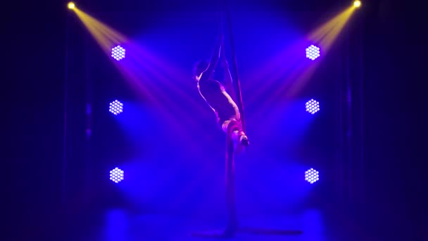En ung begåvad cirkuskvinnlig artist utför balansering på flygsilke. Akrobatisk sko i en mörk studio med blått neonljus. Långsamma rörelser. — Stockvideo
