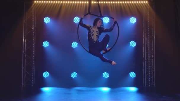 Woman aerialist performs acrobatic elements and spins in hanging aerial hoop against background of blue neon lights. — Stock Video