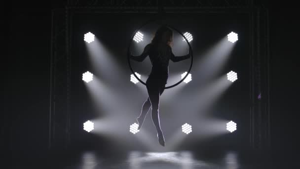 Beautiful curly aerial gymnast performs elements on aerial hoop. The graceful silhouette of a young girl in the spotlight looms out of the darkness. — Stock Video