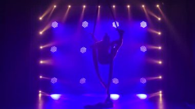 Kadın hava dengesi jimnastikçisi, mavi sahne ışıklandırmasıyla karanlık bir stüdyoda hava ipeği üzerinde dengede duruyor. İnce bir vücudun silueti. Kapat..
