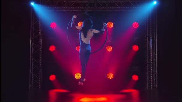 Воздушная гимнастка выполняет акробатические трюки на воздушном обруче. Женщина делает несколько акробатических элементов на воздушном обруче в помещении. Акробатическое шоу в темной студии с красной подсветкой сцены. — стоковое видео
