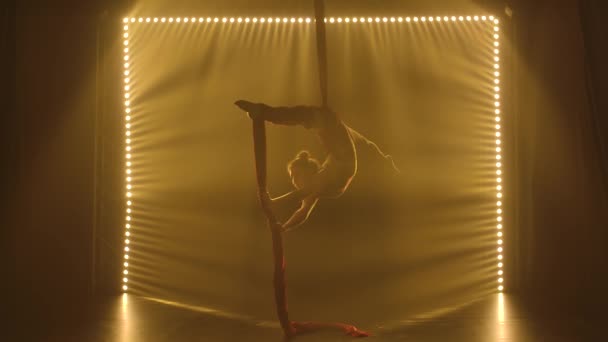 Silueta joven mujer realiza los elementos acrobáticos en la seda del aire. Aerialista está girando en el aire contra el telón de fondo de la luz suave puesta en escena. — Vídeos de Stock