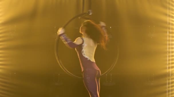 En kvinnlig akrobat roterar i en luftbåge och utför trick. Spännande akrobatisk show i en mörk studio med gult scenljus. Närbild. — Stockvideo