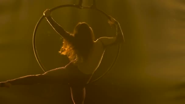 Silhouette female acrobat rotates in an air hoop and performs tricks. Exciting acrobatic show in a dark studio with yellow stage lighting. Close up. — Stock Video