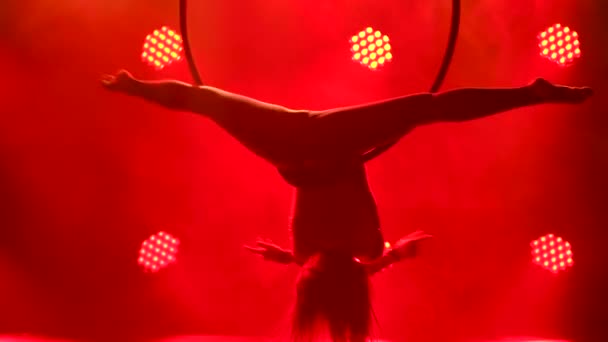 A young woman performs the acrobatic elements in the air ring. Aerial acrobat in a dark studio with red stage lighting. Silhouette close up. — Stock Video