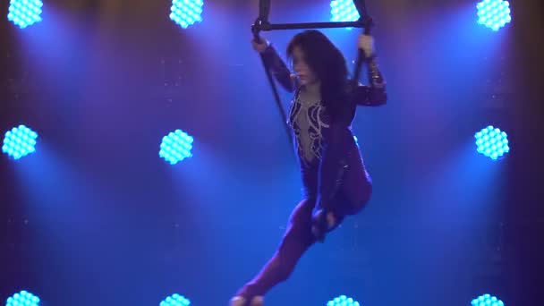Flexible woman hanging in an aerial acrobatics ring performs complex tricks. Aerial acrobat in a dark studio with blue stage lighting. Close up. — Stock Video