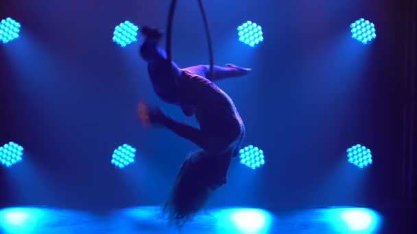 Silhouette of flexible woman hanging in an aerial acrobatics ring performs complex tricks. Aerial acrobat close up in a dark studio with blue stage lighting. — Stock Video