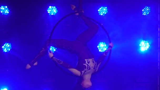 Flexibilní žena zavěšená ve vzdušném akrobacickém kruhu provádí složité triky. Letecký akrobat v tmavém ateliéru s modrým osvětlením jeviště. Zavřít. — Stock video