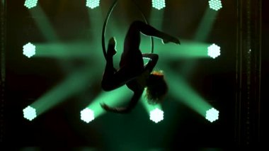 Hava jimnastiğinin silueti hava çemberinde akrobatik numaralar sergiliyor. Karanlık bir stüdyoda yeşil sahne ışıklandırmasıyla heyecan verici bir akrobatik gösteri. Kapat..