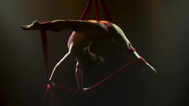 Hava ipeği üzerinde genç bir kadın oyuncunun akrobatik performansı. Bir kadının silueti, arka plan ışıklarıyla havada dönüyor. Kapat..