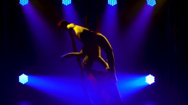 Genç bir kadın hava ipeğiyle akrobatik numaralar yapıyor. Mavi sahne ışıklandırması olan karanlık bir stüdyoda heyecan verici bir akrobatik gösteri. İnce bir vücudun silueti. Kapat..