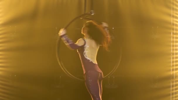 A female acrobat rotates in an air hoop and performs tricks. Exciting acrobatic show in a dark studio with yellow stage lighting. Close up. — Stock Video