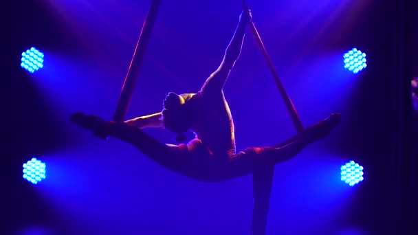 Młoda kobieta robi akrobatyczne sztuczki z powietrznym jedwabiem. Ekscytujący akrobatyczny show w ciemnym studio z niebieskim oświetleniem sceny. Sylwetka szczupłego ciała. Zamknij się.. — Wideo stockowe