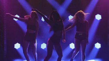 Three sensual long haired woman shaking her ass dancing twerk in studio against a background of blue lights. Silhouette. Close up. Slow motion.