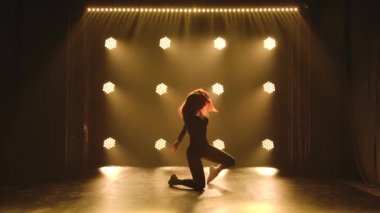 Kızıl saçlı, siyah taytlı, genç seksi bir kadın karanlık bir stüdyoda dans ediyor. Sahne ışığının arka planında siluetler var. Yavaş çekim.