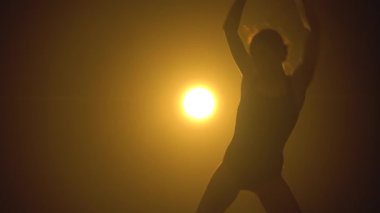 Siluet ince, esnek, siyah elbiseli bir balerin stüdyonun sarı sahne ışıkları altında karanlıkta zarif bir şekilde dans ediyor..