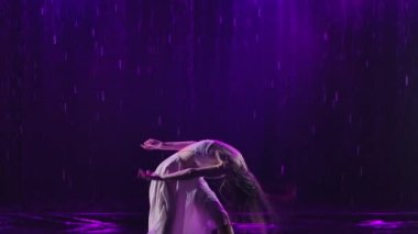 Islak elbiseli güzel bir kadın yağmur damlalarının altında tutkuyla dans ediyor. Stüdyo ışığı olan karanlık bir arka plan. Ağır çekim. Kapat..