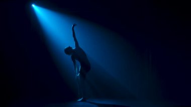 Siluet, esnek, siyah elbiseli genç bir balerin stüdyonun mavi sahne ışıkları altında karanlıkta zarif bir şekilde dans ediyor. Yavaş çekim.